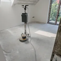 Szlifowanie posadzki w nowym, jasnym pomieszczeniu z oknem. Na podłodze stoi maszyna do szlifowania. W tle widoczne ściany przygotowane do malowania.