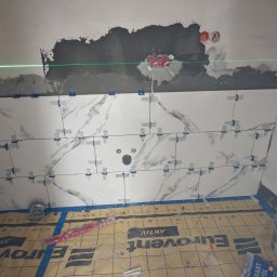 Kamil Gałka - Białe płytki imitujące marmur, układane z użyciem krzyżyków dystansowych, poziomowane laserowo. Podłoga zabezpieczona folią budowlaną. Widoczne instalacje elektryczne.