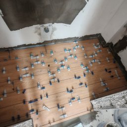 Kamil Gałka - Układanie drewnopodobnych płytek na podłodze w łazience, z użyciem klipsów poziomujących. Widoczne niedokończone ściany i sufit w surowym stanie.