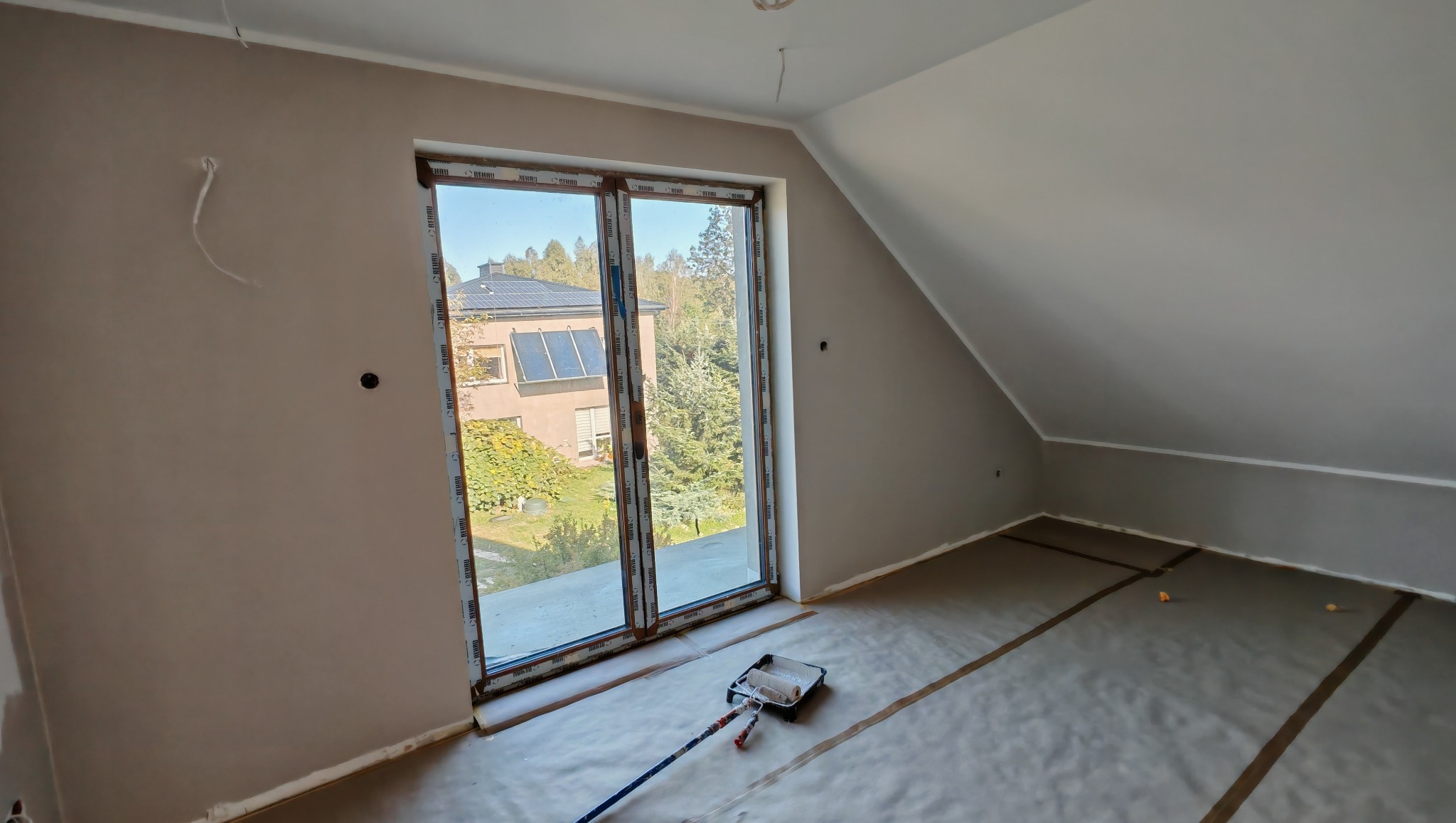 Poddasze po malowaniu na beżowo, widok na okno z widokiem na dom i zieleń. Na podłodze folia ochronna i wałek malarski. Ściany gładkie, świeżo pomalowane.