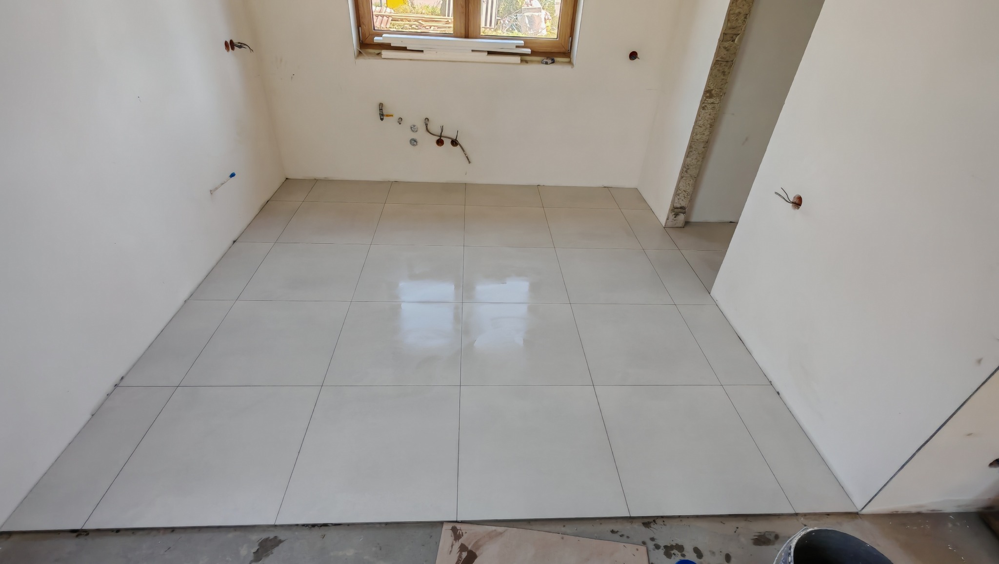 Jasne, kwadratowe płytki podłogowe w świeżo wyremontowanym, pustym pomieszczeniu z oknem. Widoczne wystające kable i rury instalacyjne na ścianach.