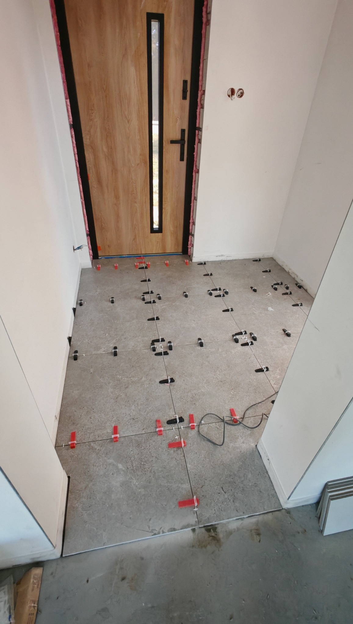 Układanie płytek podłogowych w korytarzu z użyciem systemu poziomowania, widoczna drewniana okleina drzwi wejściowych i białe ściany.