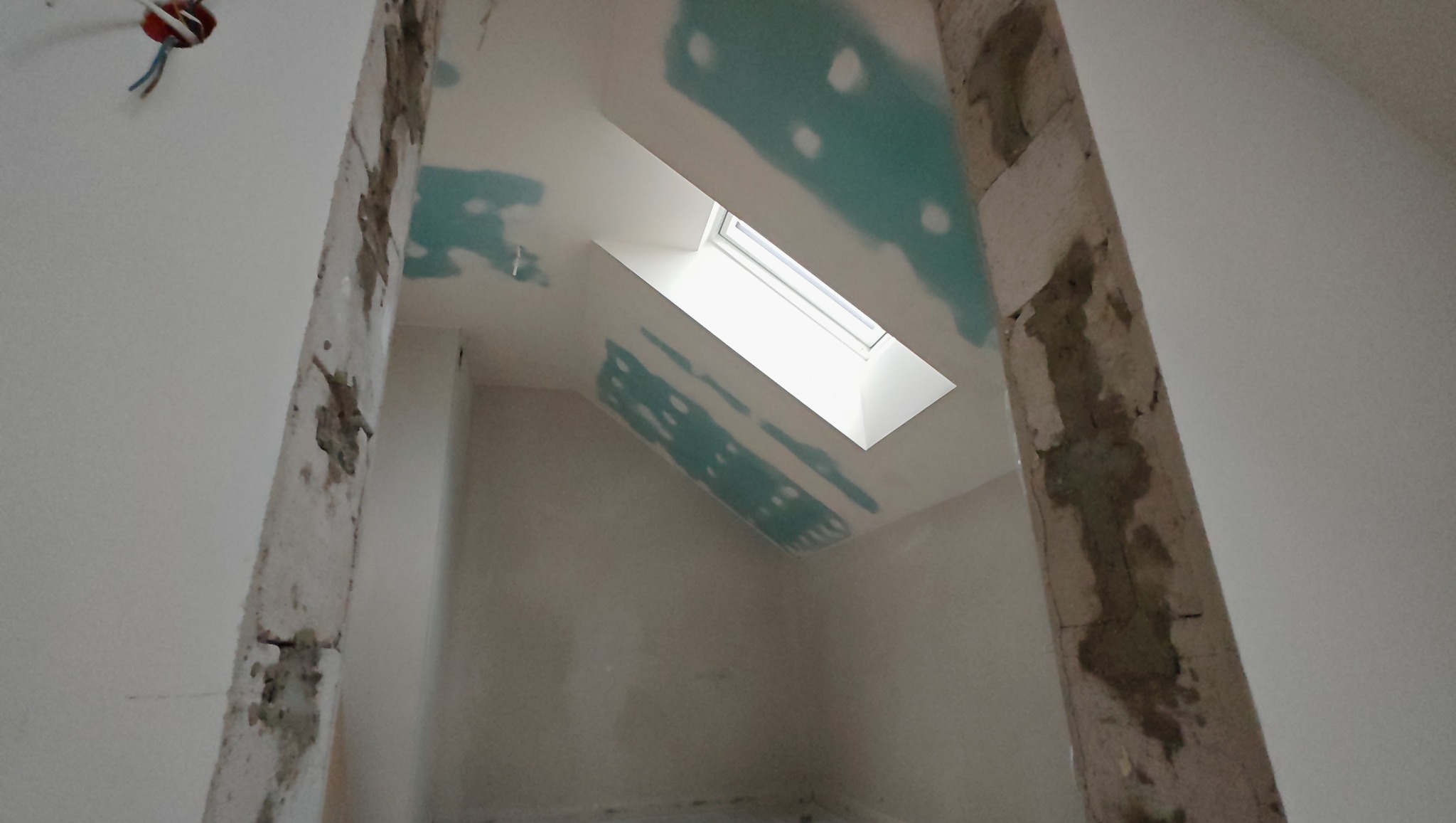 Poddasze w trakcie prac wykończeniowych: szpachlowanie ścian i sufitu, widoczne okno dachowe. Fragment instalacji elektrycznej przy suficie. Surowy stan framugi.