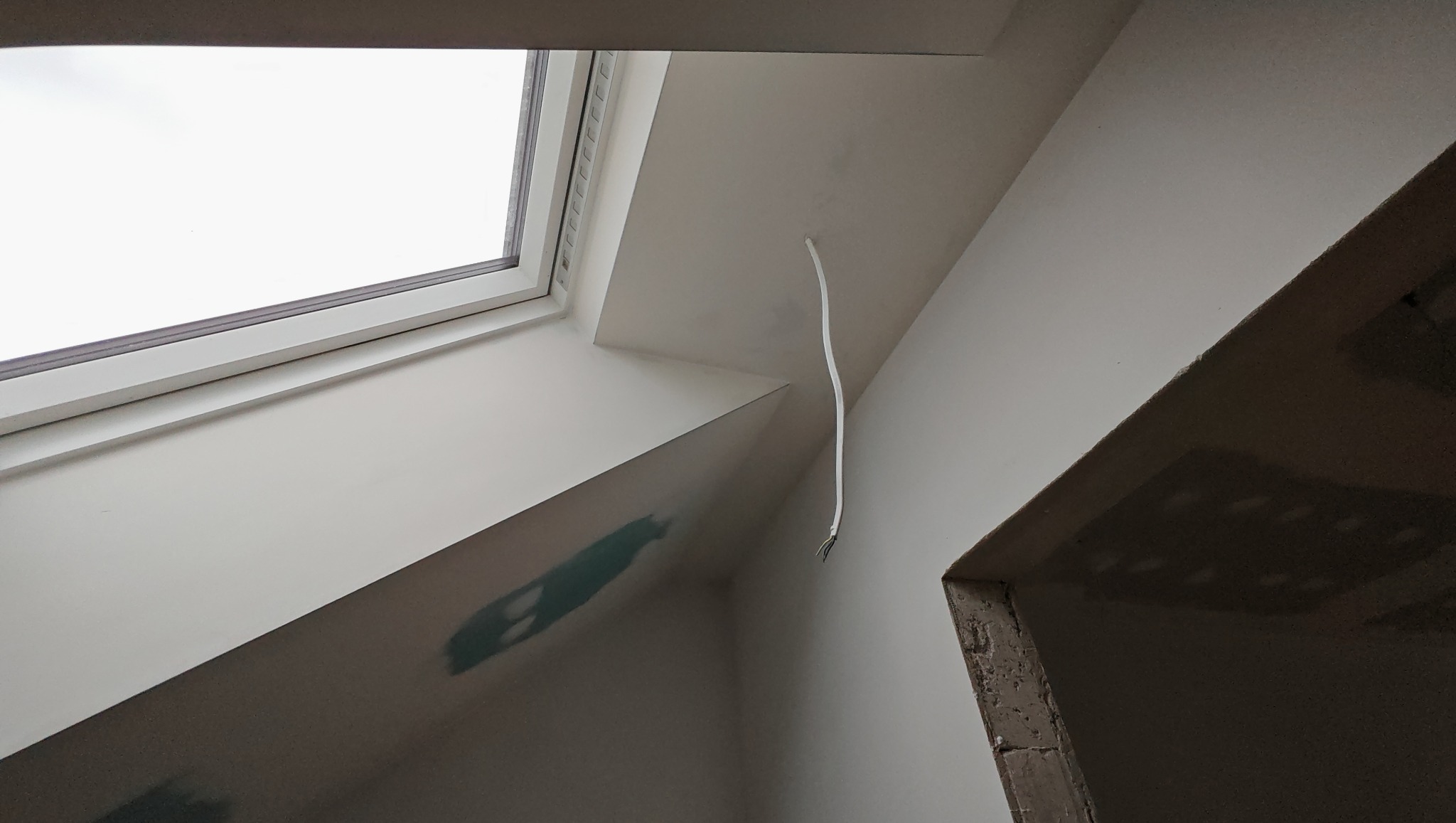 Poddasze z oknem dachowym w trakcie wykończenia. Widoczny kabel elektryczny wystający ze ściany i surowy otwór drzwiowy. Ściany pomalowane na biało.