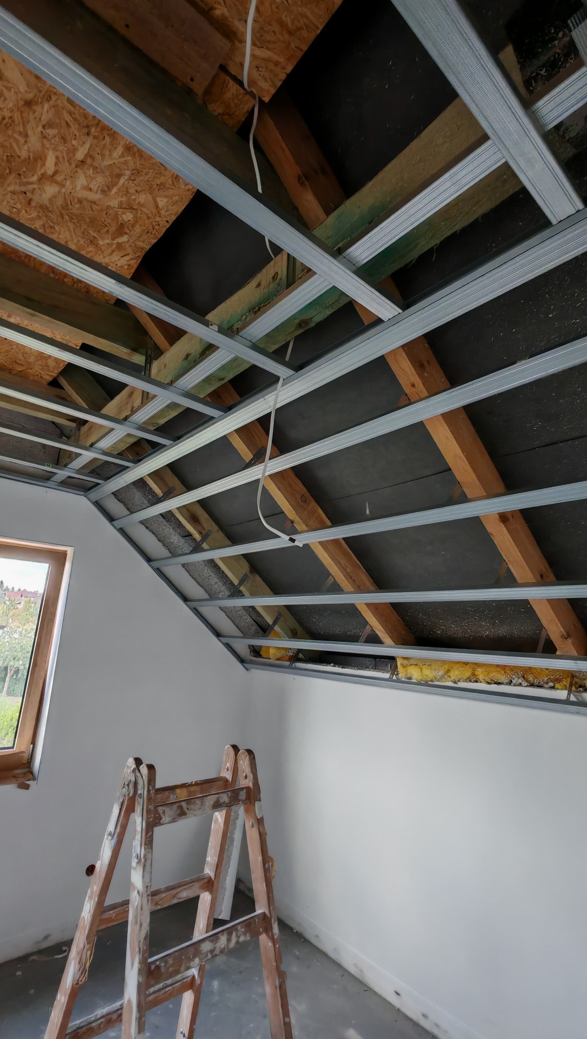 Sufit podwieszany z metalowym stelażem i izolacją, widok z dołu. Drewniana drabina oparta o ścianę, okno w tle. Prace wykończeniowe poddasza.