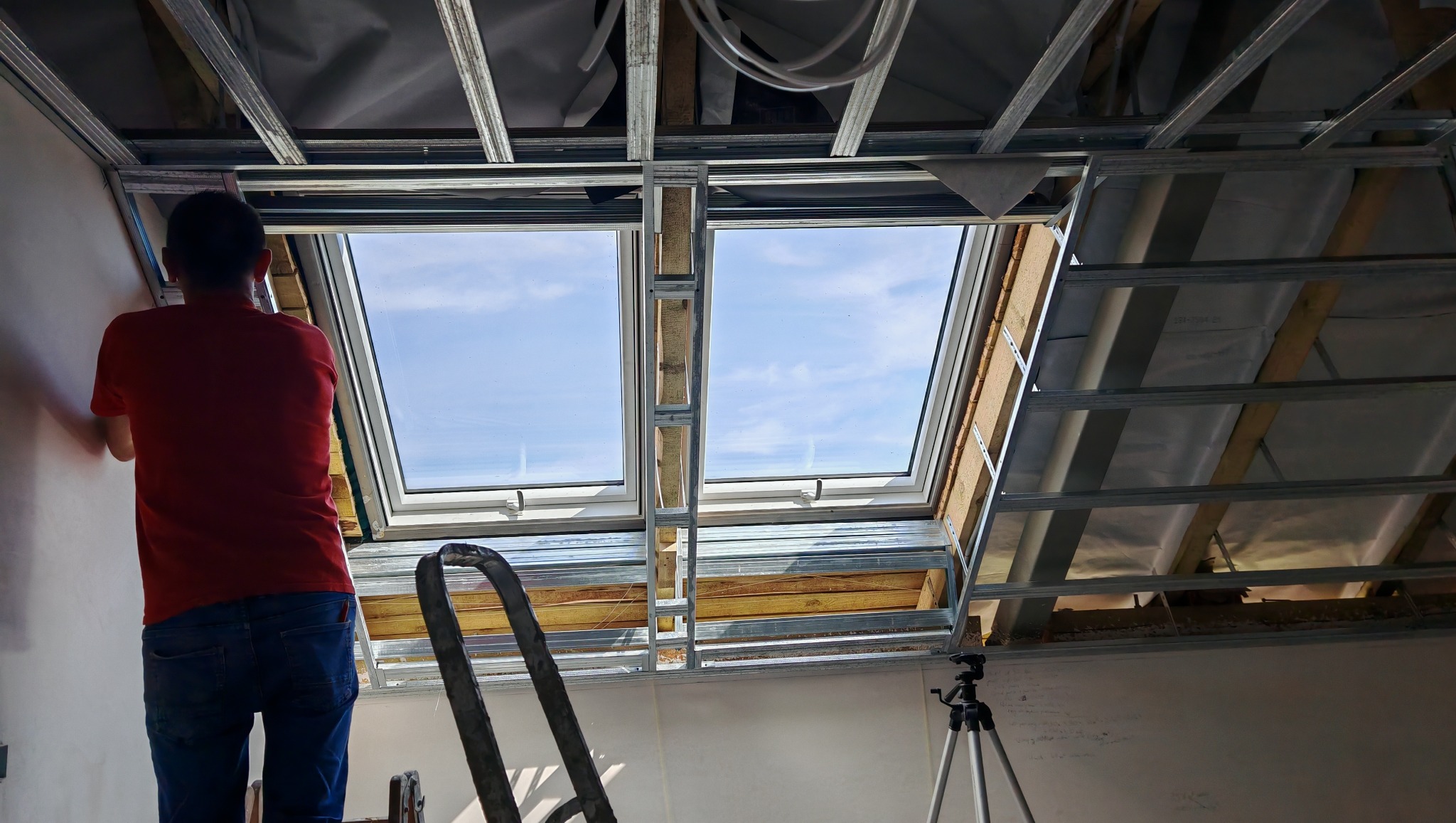 Montaż stelaża pod płytę G-K na poddaszu z oknami dachowymi. Widoczny pracownik w czerwonej koszulce i drabina. W tle niebo przez okna.