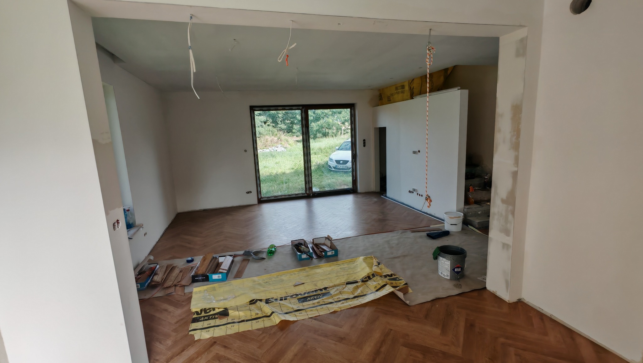 Salon w trakcie remontu: parkiet w jodełkę, częściowo ułożony, widoczne narzędzia i materiały budowlane, okno balkonowe z widokiem na zieleń, ściany przygotowane do malowania.