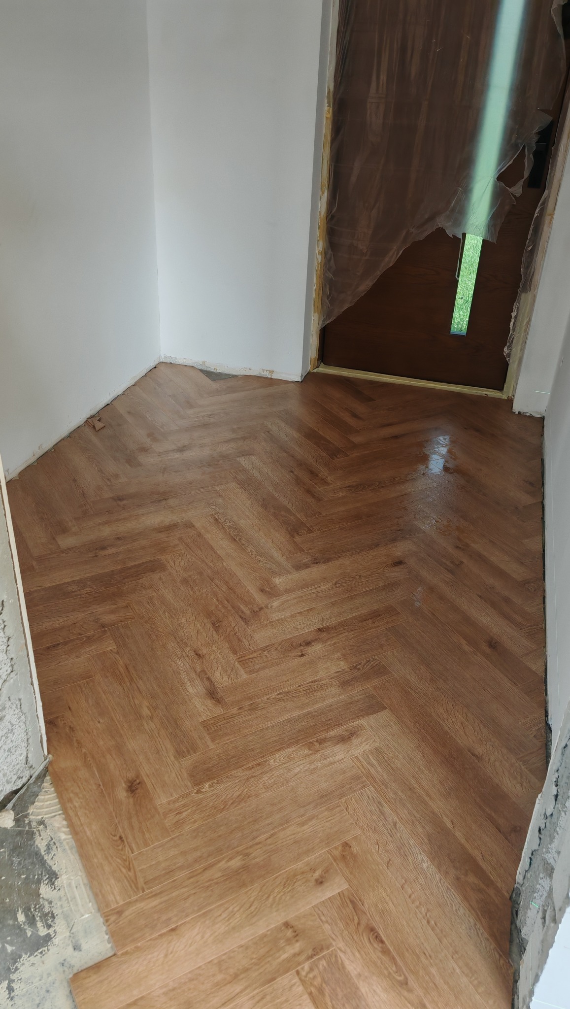 Ułożona podłoga z paneli w jodełkę, widoczna przy białej ścianie i zabezpieczonych folią drzwiach, częściowo wykończona przy listwach.