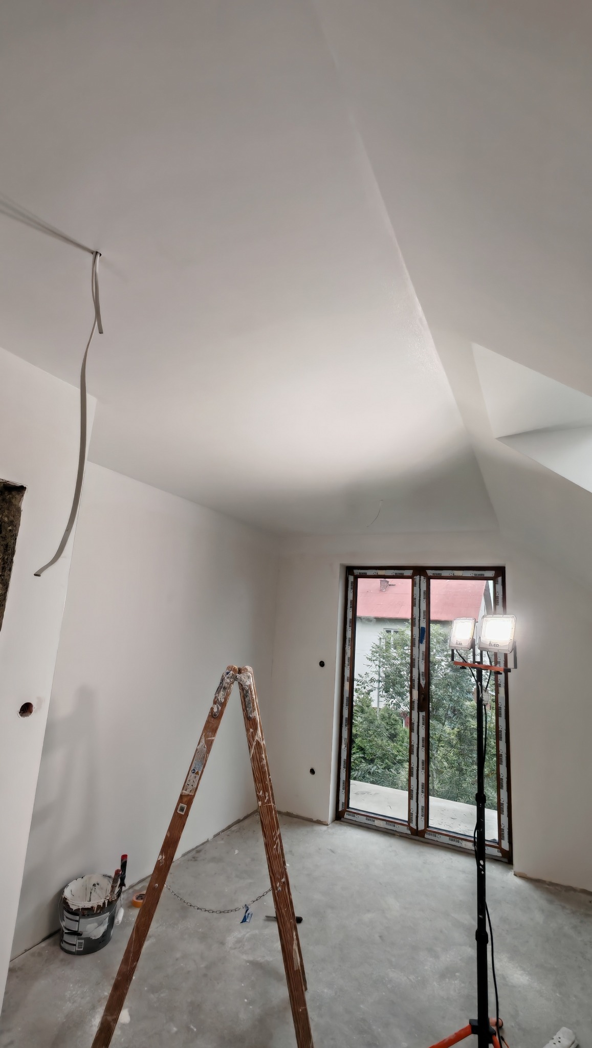 Poddasze po malowaniu na biało, widoczna drabina, wiadro z farbą, lampa oświetleniowa i okno. Świeżo pomalowane ściany na biało.
