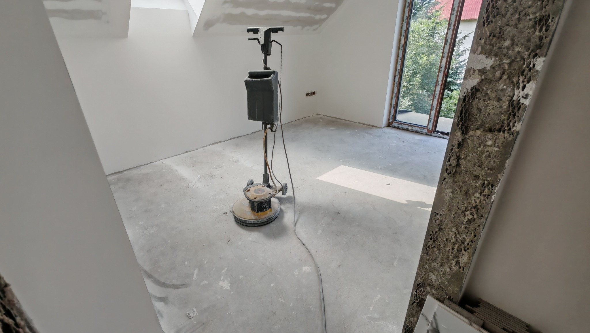 Szlifowanie posadzki w nowym, jasnym pomieszczeniu z oknem. Na podłodze stoi maszyna do szlifowania. W tle widoczne ściany przygotowane do malowania.