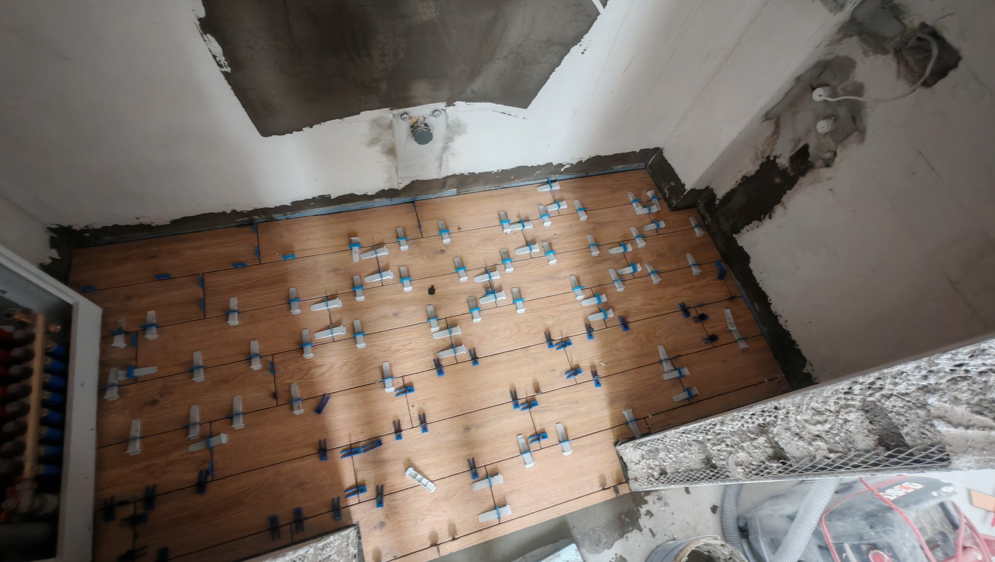 Układanie drewnopodobnych płytek na podłodze w łazience, z użyciem klipsów poziomujących. Widoczne niedokończone ściany i sufit w surowym stanie.