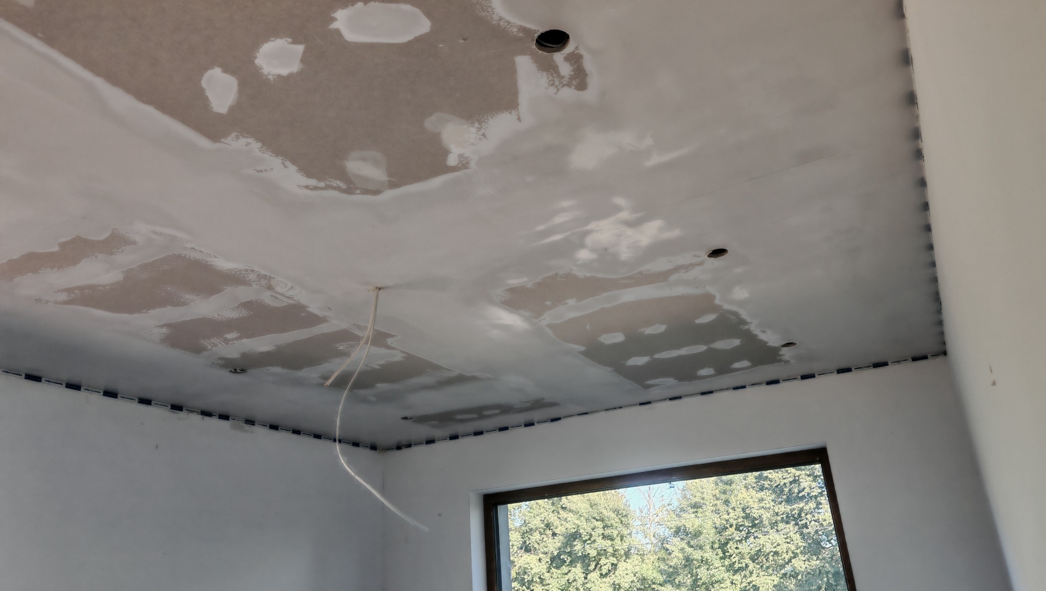 Płyta karton-gips na suficie w trakcie szpachlowania, widoczne łączenia i otwory na oświetlenie. W tle okno z widokiem na zieleń.