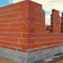 Wznoszenie muru z czerwonych, porowatych pustaków ceramicznych na fundamencie z szarego betonu. Widoczne niebo i inne elementy konstrukcji w tle.