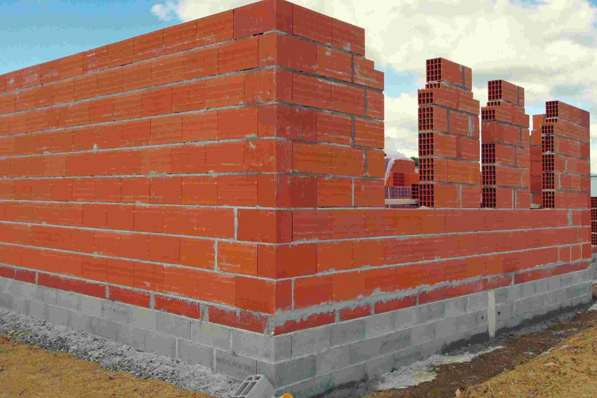 Wznoszenie muru z czerwonych, porowatych pustaków ceramicznych na fundamencie z szarego betonu. Widoczne niebo i inne elementy konstrukcji w tle.