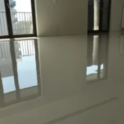 Jasna, połyskująca posadzka w pustym pokoju z dużymi oknami balkonowymi. Widoczne odbicia okien w gładkiej powierzchni podłogi.