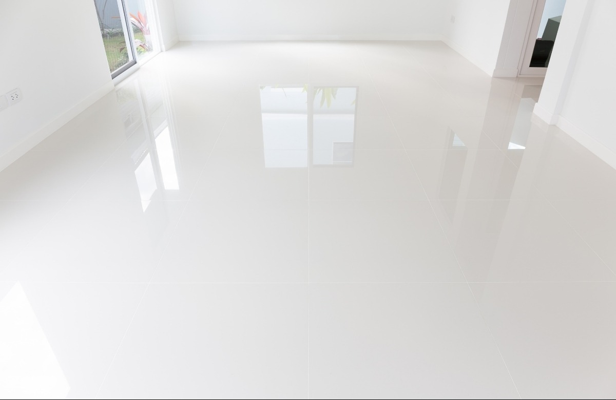 Błyszcząca, jasna posadzka w minimalistycznym wnętrzu z oknem i drzwiami. Powierzchnia odbija światło, tworząc efekt czystości i przestronności. Widoczne subtelne fugi.