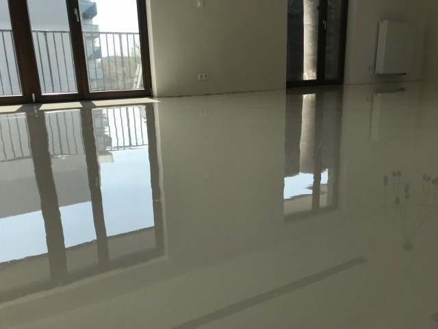 Jasnoszara, błyszcząca posadzka w nowym mieszkaniu z dużymi oknami balkonowymi, odbijającymi światło. Minimalistyczny design, widoczne instalacje elektryczne na ścianie.