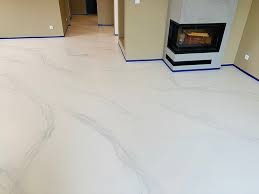 Biała posadzka z efektem marmuru w salonie z kominkiem, zabezpieczona niebieską taśmą malarską przy ścianach. Widok z góry, neutralne oświetlenie.