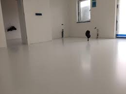 Błyszcząca, jasnoszara posadzka w minimalistycznym wnętrzu z białymi ścianami i oknem. Widoczne pionowe rury przy ścianie. Gładka powierzchnia odbija światło.