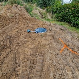 Deka Przydomowe oczyszczalnie ścieków - Zamknięty właz przydomowej oczyszczalni ścieków na nierównym terenie, z widocznymi śladami opon i pomarańczowym oznaczeniem na ziemi.