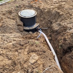 Deka Przydomowe oczyszczalnie ścieków - Montaż zbiornika z rurą w wykopie. Widoczne ślady prac ziemnych i drabina w tle. Instalacja w trakcie realizacji, z ziemią wokół.