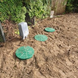 Deka Przydomowe oczyszczalnie ścieków - Instalacja przydomowej oczyszczalni: trzy zielone włazy inspekcyjne i szara skrzynka sterująca na ziemi, widok z góry na tle zieleni i ogrodzenia.