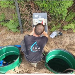 Deka Przydomowe oczyszczalnie ścieków - Montaż przydomowej oczyszczalni ścieków: instalator podłącza sterownik, widoczne dwa zielone zbiorniki i ogrodzenie z siatki.