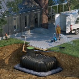 Deka Przydomowe oczyszczalnie ścieków - Czarny zbiornik retencyjny w ziemi, z podłączeniem do węża ogrodowego. W tle dom z ogrodem i pracownik z wężem. Prezentacja instalacji zbiornika na wodę deszczową.