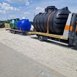 Deka Przydomowe oczyszczalnie ścieków - Transport zbiorników: czarny, niebieski i zielone, zabezpieczone pasami na lawecie samochodu dostawczego. Widoczne niebo i drzewa w tle.