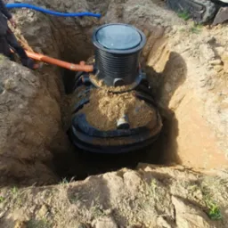 Montaż czarnego zbiornika retencyjnego w wykopie ziemnym. Widoczne rury podłączeniowe i mała koparka. Ujęcie z góry, z widocznymi butami fotografa.