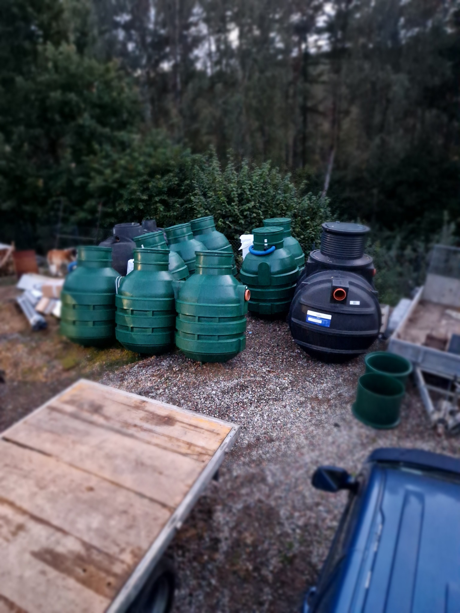 Zielone i czarne zbiorniki na żwirze, prawdopodobnie elementy instalacji sanitarnej, z przyczepką i fragmentem niebieskiego auta w kadrze.