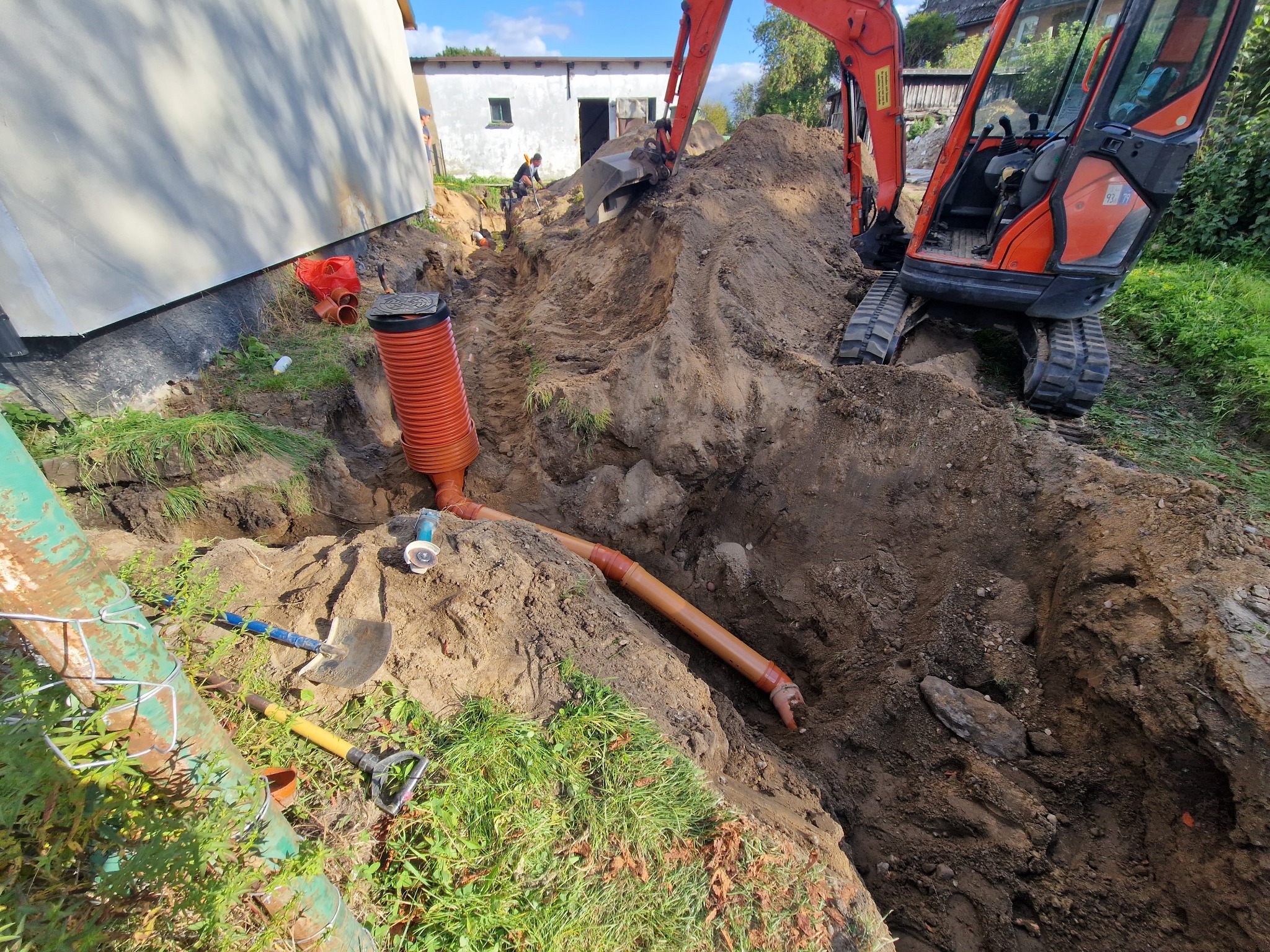 Wykop pod rury kanalizacyjne, pomarańczowe rury w ziemi, koparka w tle. Prace ziemne przy budynku, widoczne narzędzia i pracownik.