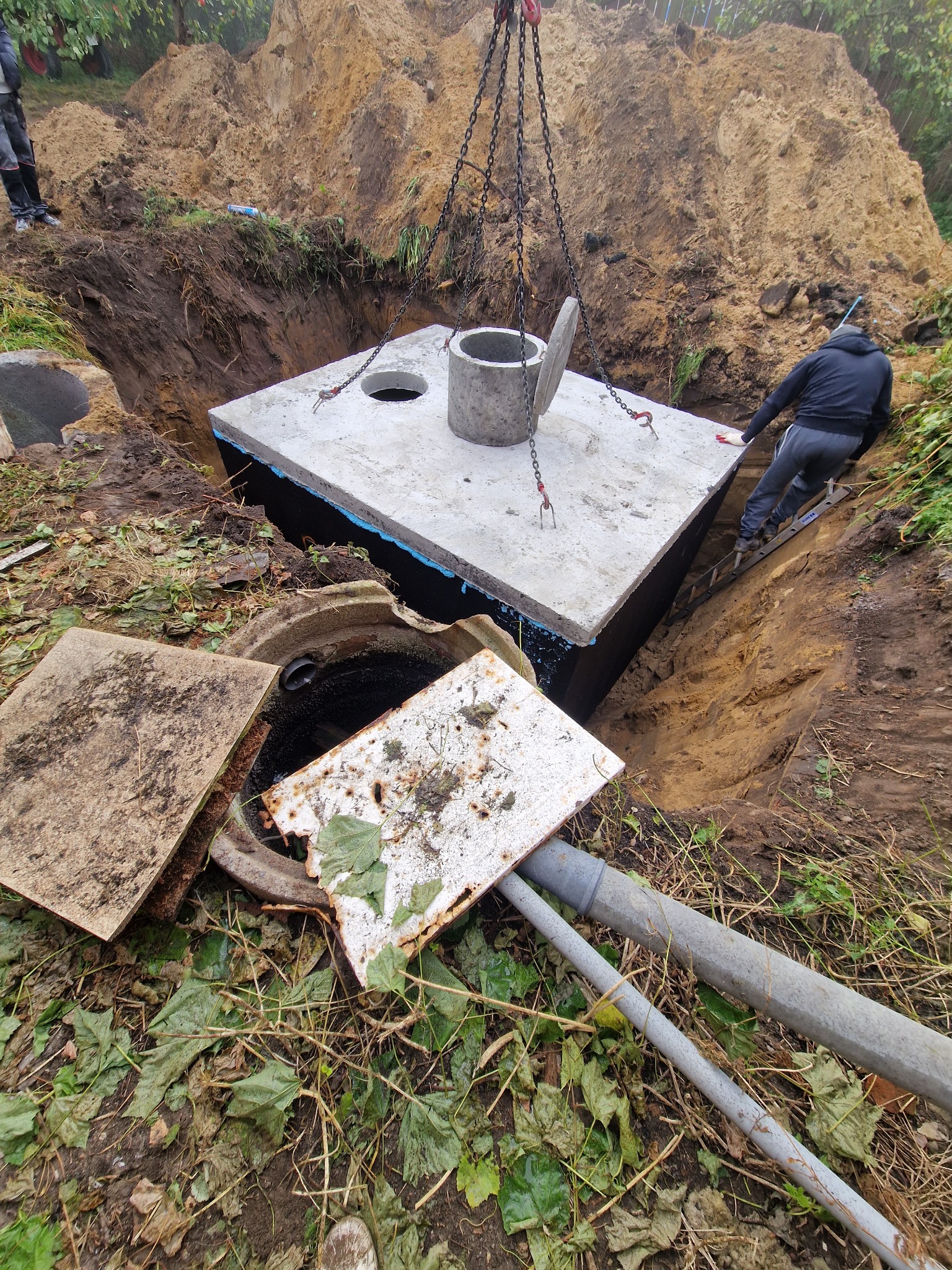 Montaż betonowego szamba w wykopie, widoczne łańcuchy, drabina i osoba nadzorująca proces. Otoczenie to ziemia i roślinność.
