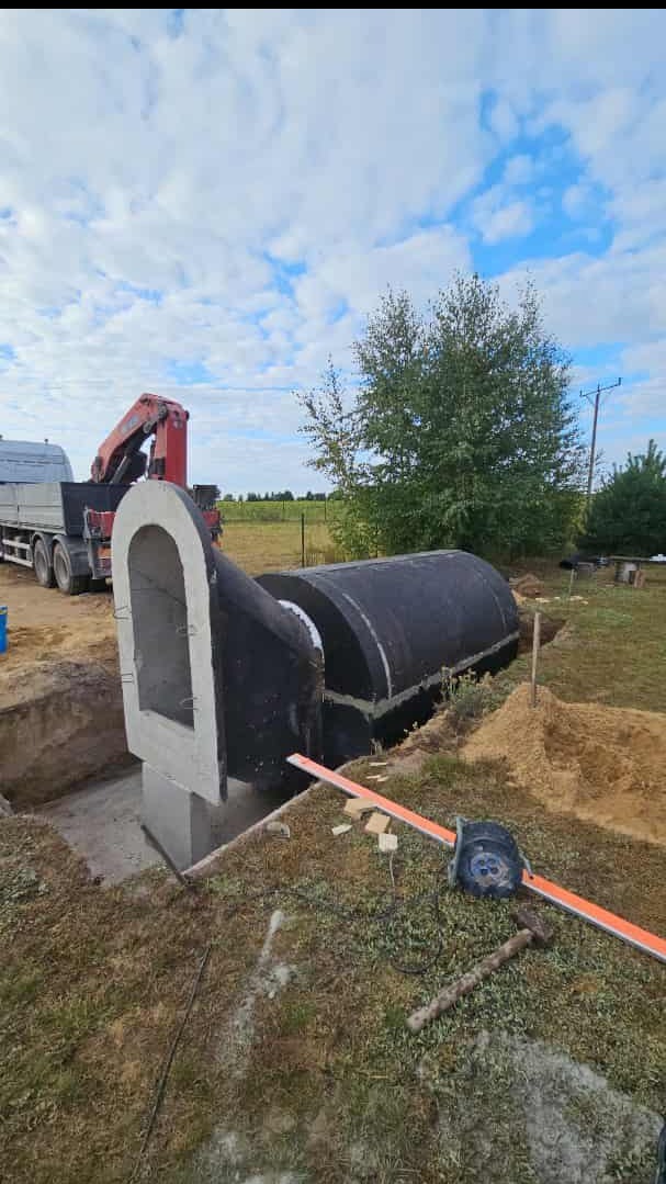 Montaż betonowego szamba: czarna, cylindryczna konstrukcja częściowo zagłębiona w ziemi, z podłączonym betonowym elementem. Widoczny dźwig i narzędzia.
