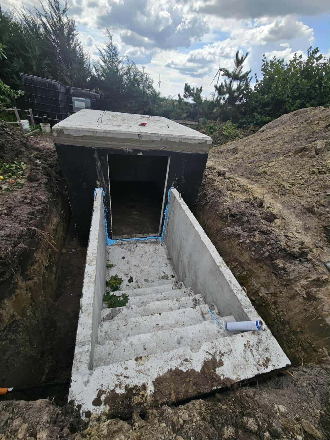 Betonowy zbiornik z wejściem schodami, wkopany w ziemię, otoczony wykopem. Widoczne uszczelnienia i elementy montażowe. Tło: drzewa i pochmurne niebo.