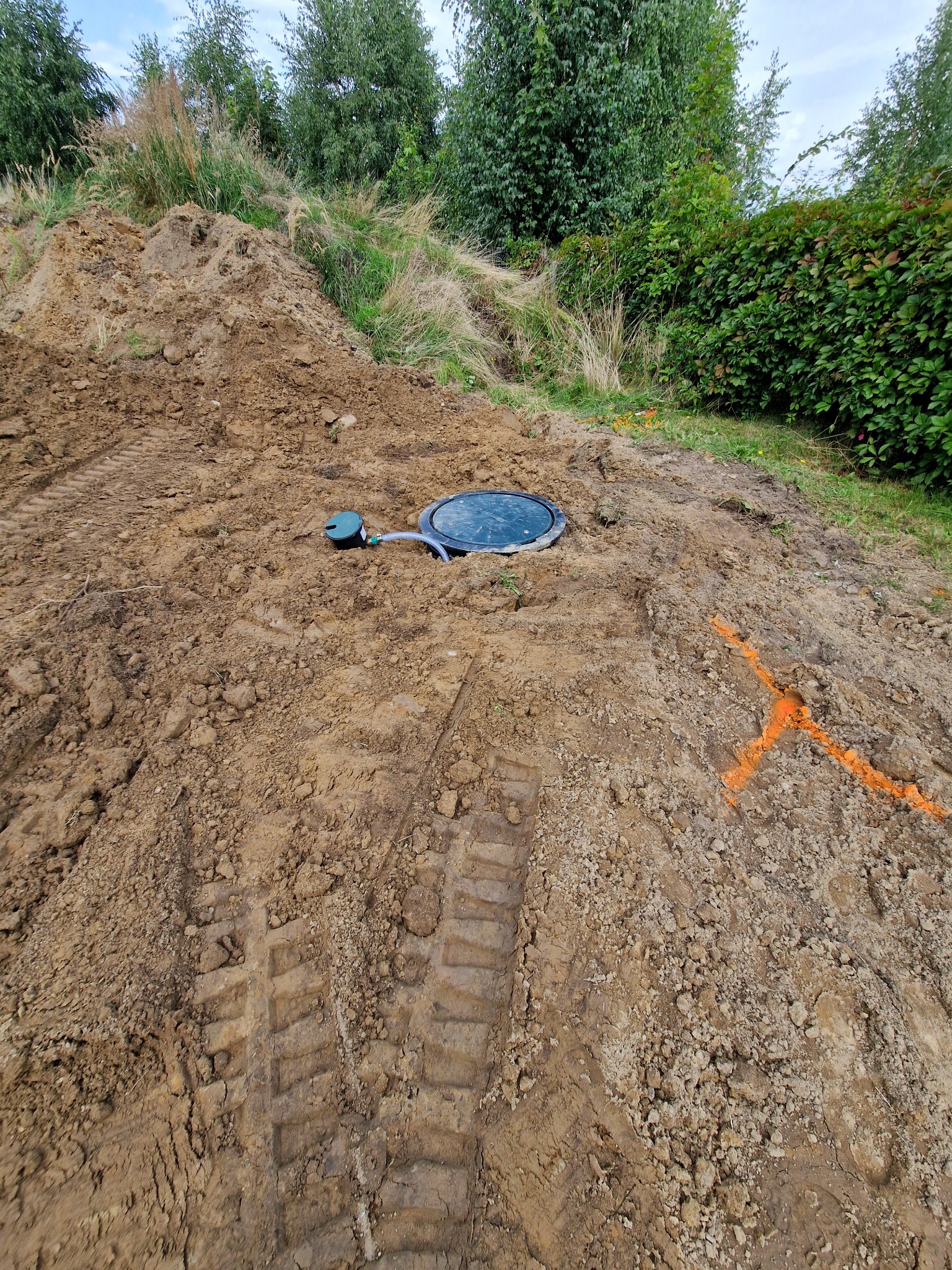 Zamknięty właz przydomowej oczyszczalni ścieków na nierównym terenie, z widocznymi śladami opon i pomarańczowym oznaczeniem na ziemi.