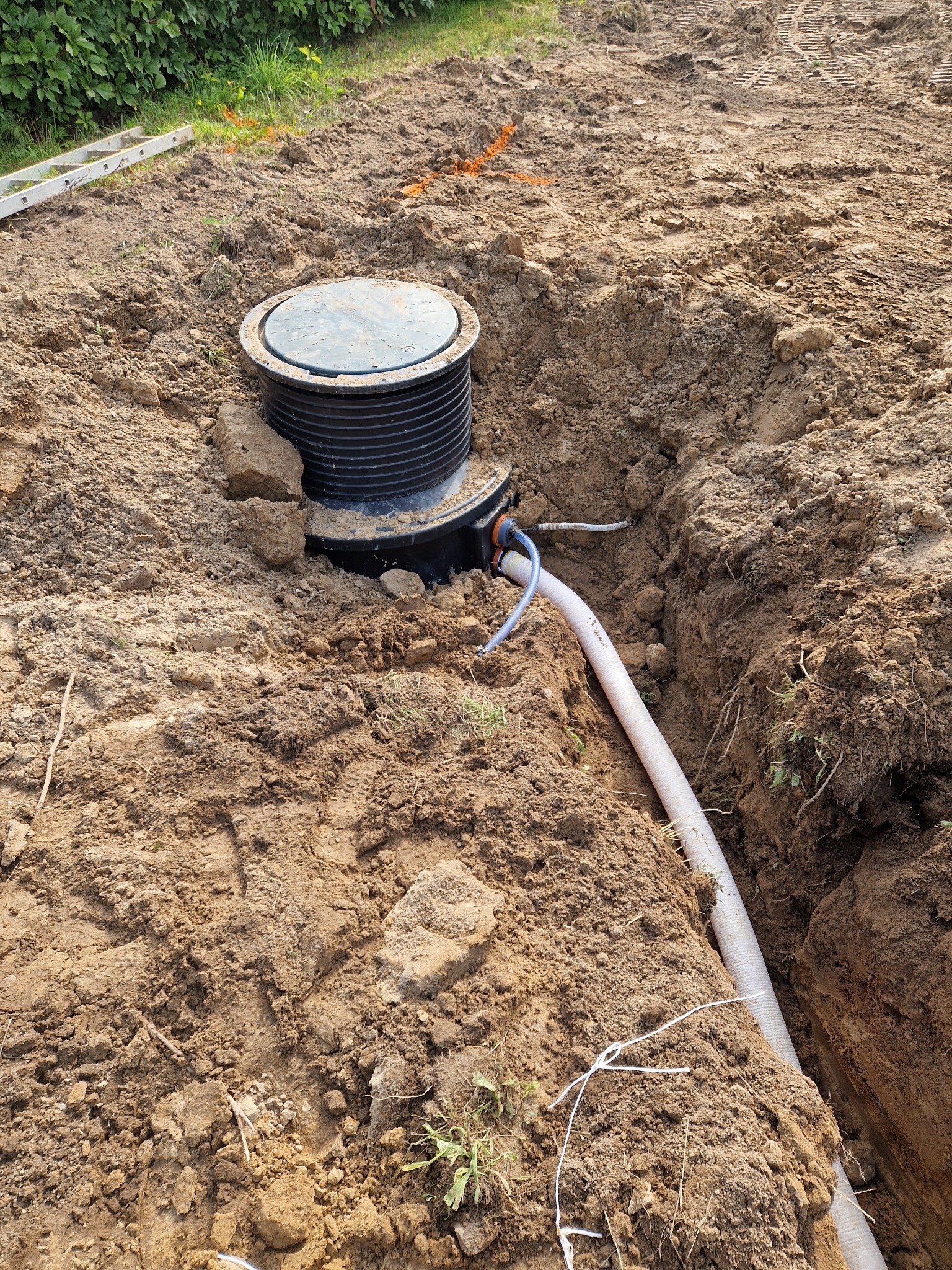 Montaż zbiornika z rurą w wykopie. Widoczne ślady prac ziemnych i drabina w tle. Instalacja w trakcie realizacji, z ziemią wokół.