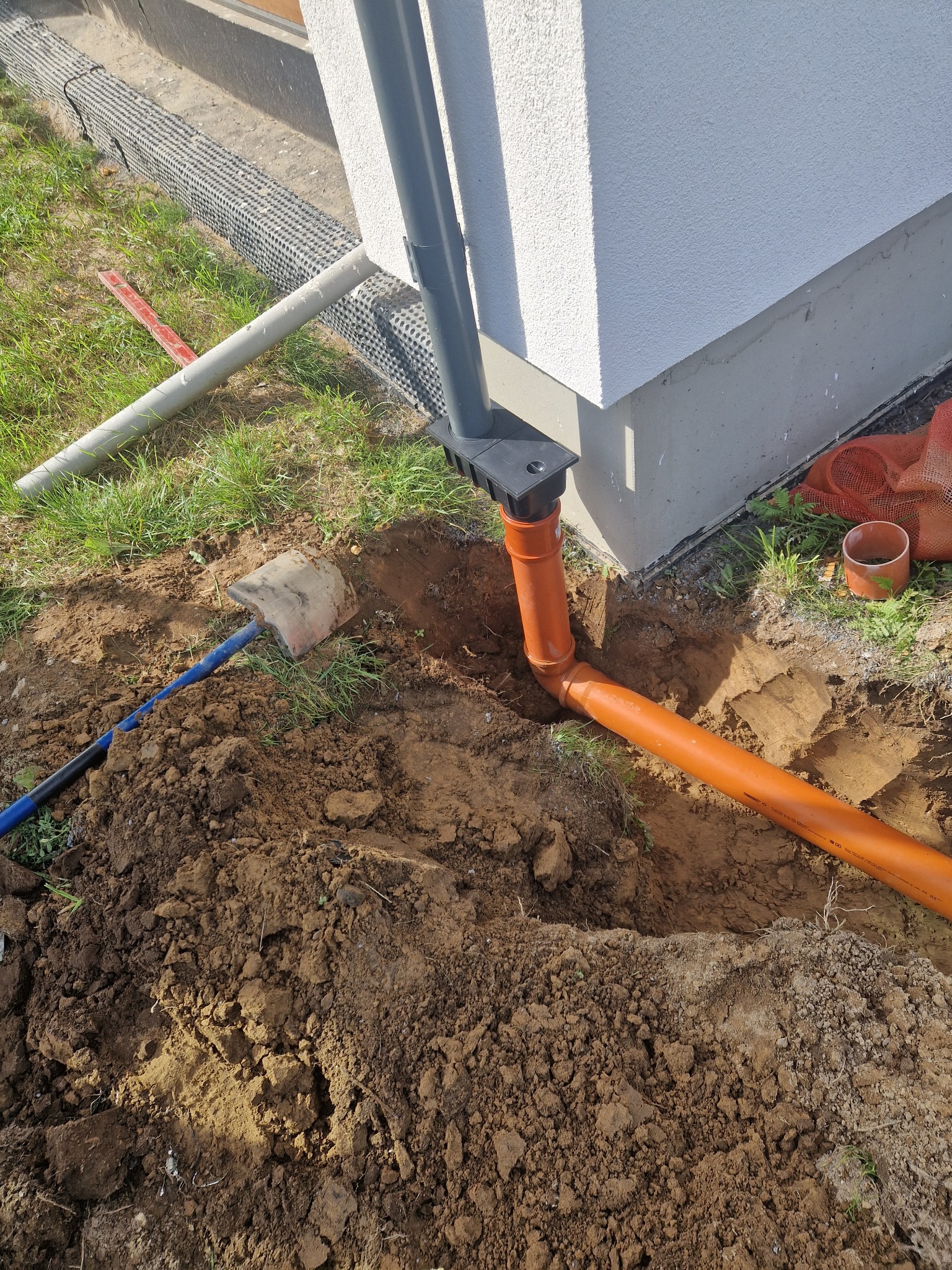 Montaż rur kanalizacyjnych pomarańczowych przy fundamencie domu. Wykopana ziemia, widoczna łopata i fragment rury spustowej.