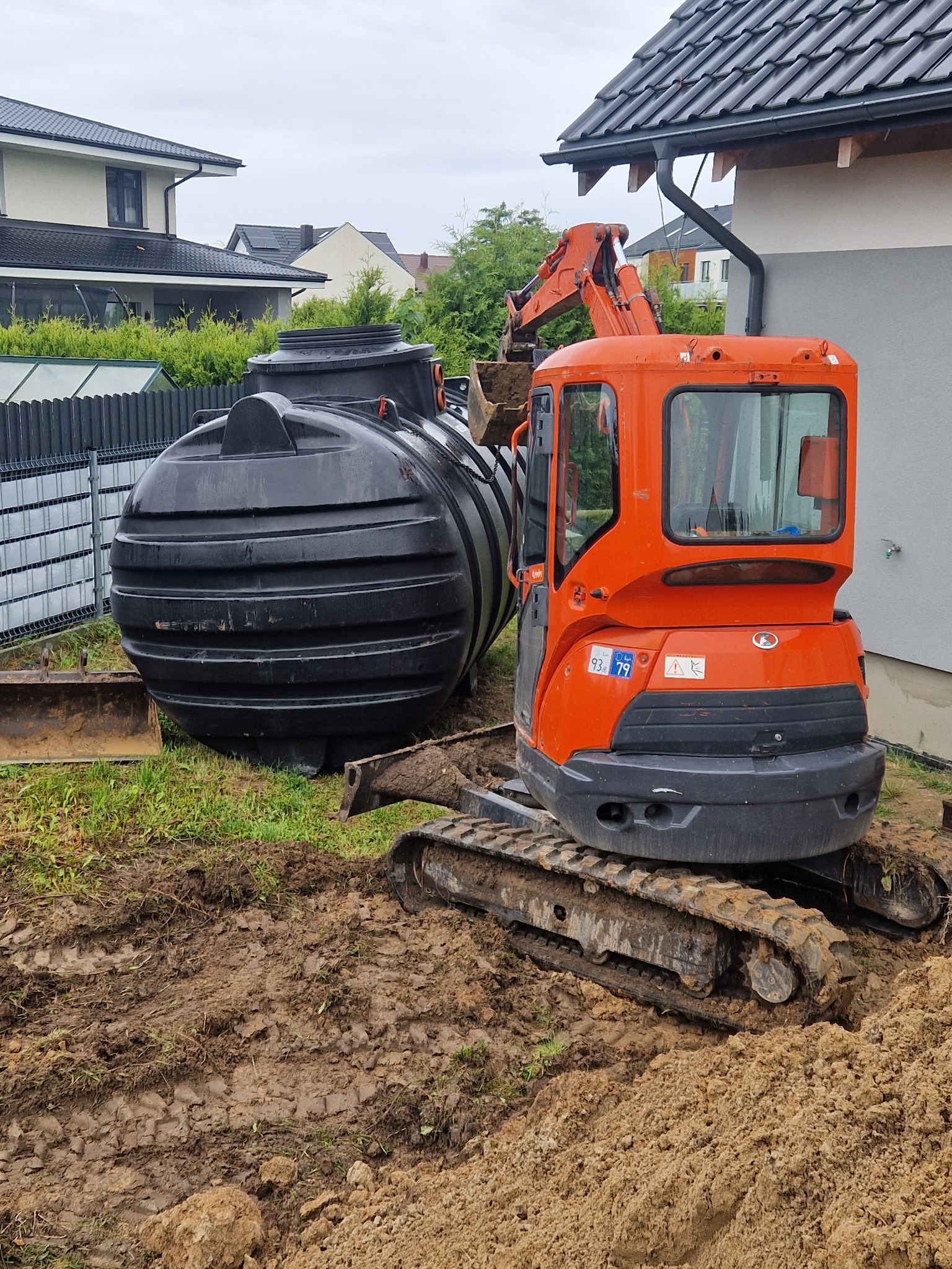 Czarny zbiornik retencyjny obok pomarańczowej mini koparki na błotnistym terenie, widok z przodu, dom w tle.