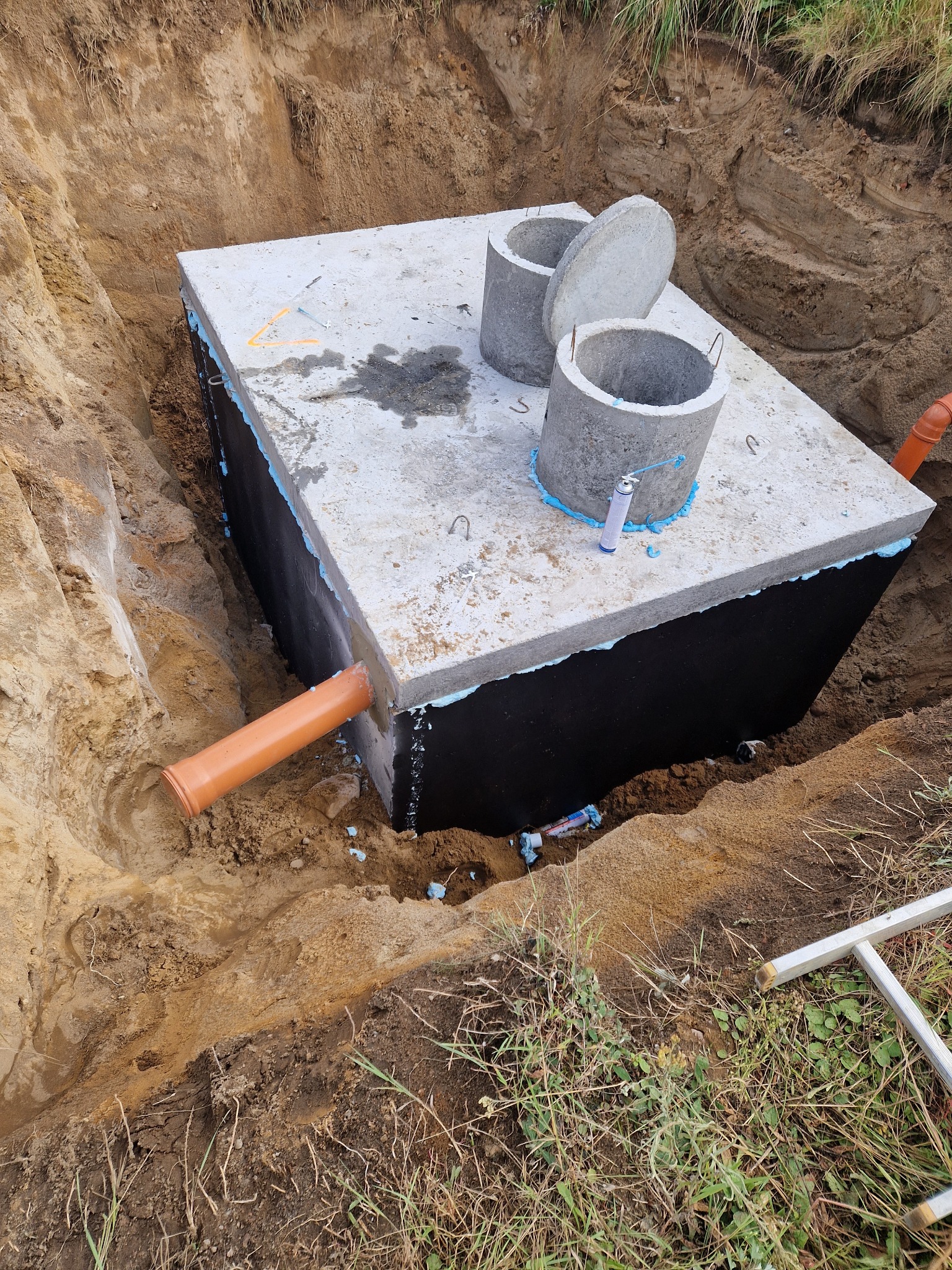 Betonowy zbiornik z rurą i dwoma kręgami w wykopie, uszczelniony pianką i czarną folią. Widoczne narzędzia i trawa. Ujęcie z góry.