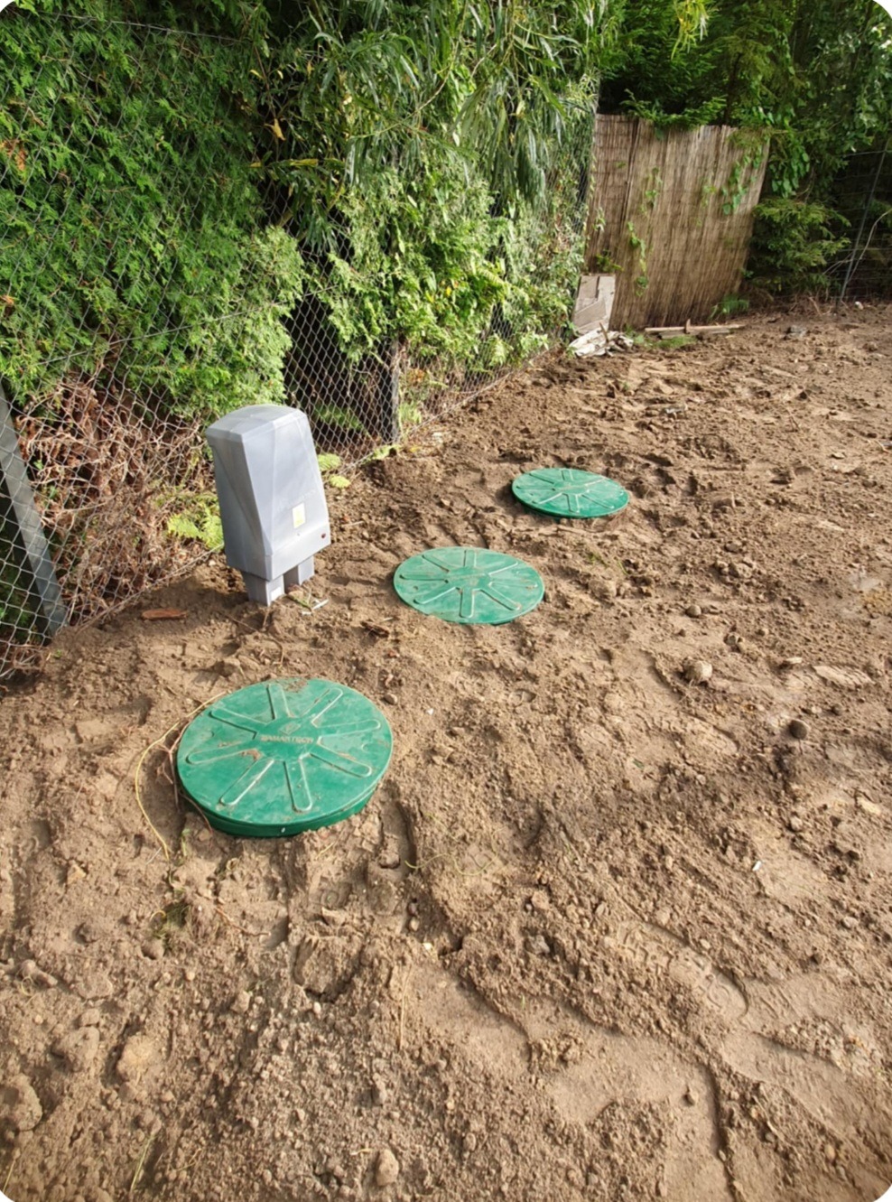 Instalacja przydomowej oczyszczalni: trzy zielone włazy inspekcyjne i szara skrzynka sterująca na ziemi, widok z góry na tle zieleni i ogrodzenia.