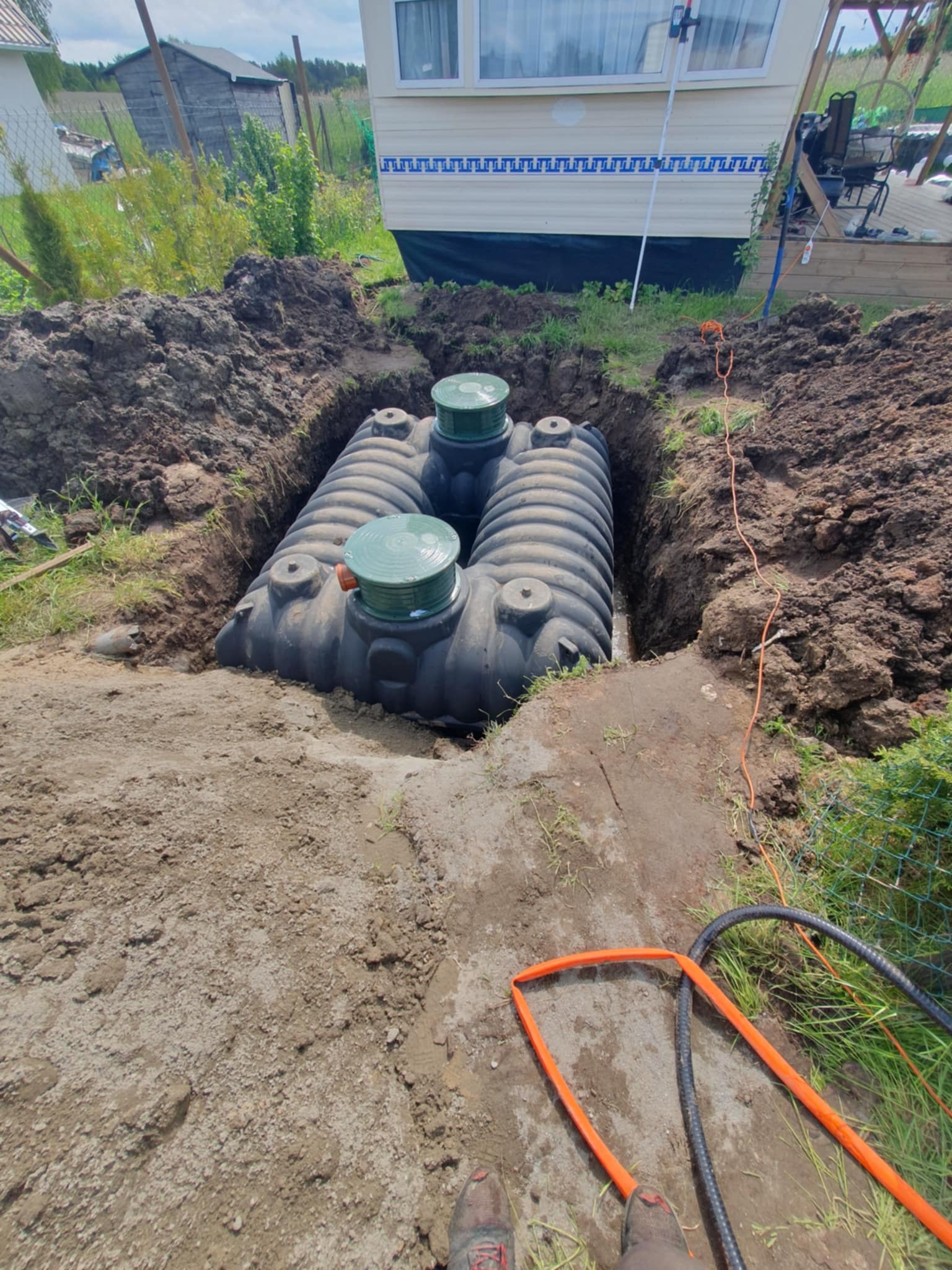 Montaż zbiornika retencyjnego w ziemi: dwa połączone zbiorniki z zielonymi pokrywami, częściowo zasypane ziemią, widoczne przewody i fragment buta w kadrze.