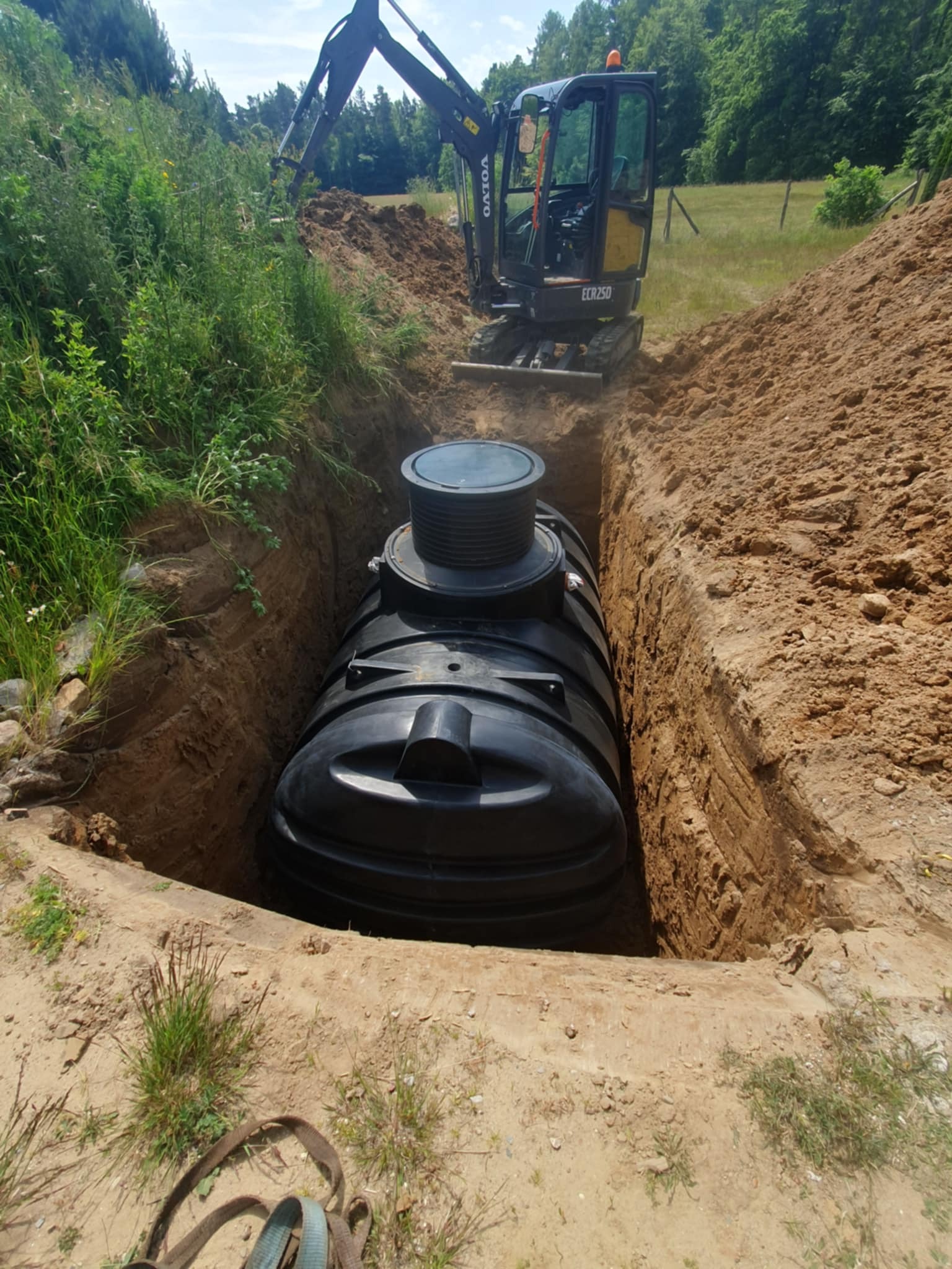 Czarny zbiornik w wykopie, obok koparka Volvo ECR25D. Widoczne ściany wykopu z piasku i trawa. Montaż zbiornika w ziemi.