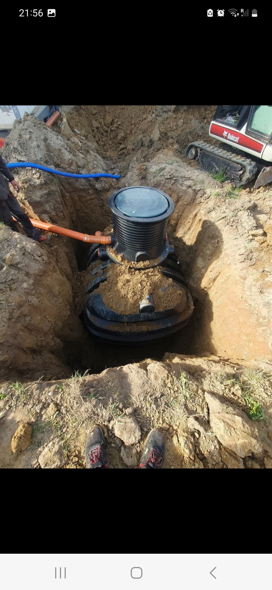 Montaż czarnego zbiornika retencyjnego w wykopie ziemnym. Widoczne rury podłączeniowe i mała koparka. Ujęcie z góry, z widocznymi butami fotografa.