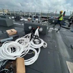 Instalacja przewodów wentylacyjnych na dachu budynku, pracownik w kasku w tle. Widoczne elementy wentylacji i przewody w rolkach. Pochmurne niebo.