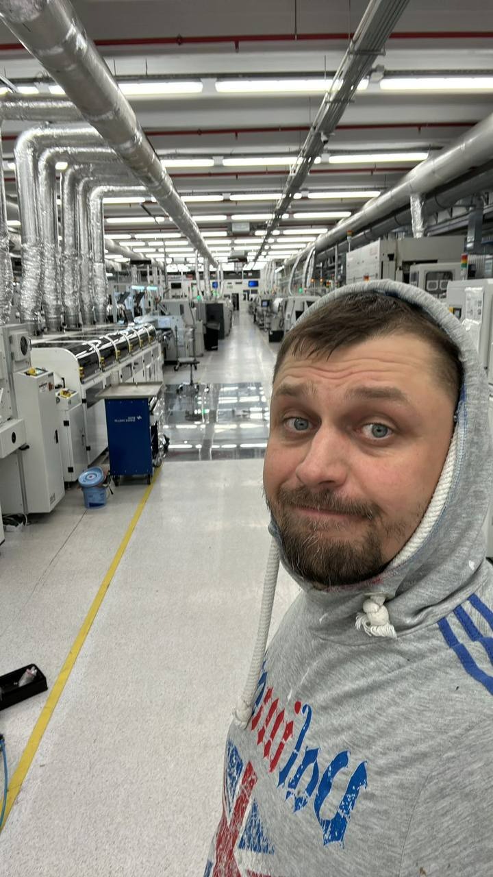 Mężczyzna w bluzie z kapturem na tle hali produkcyjnej z systemem rur wentylacyjnych pod sufitem i maszynami w tle. Ujęcie z dołu, selfie.