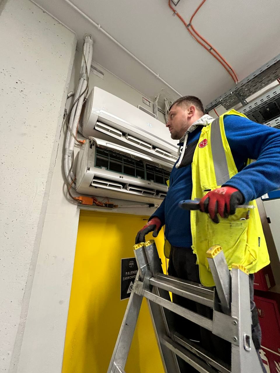 Monter w kamizelce na drabinie serwisuje klimatyzator na ścianie, widoczne przewody i instalacja. Żółte drzwi w tle.