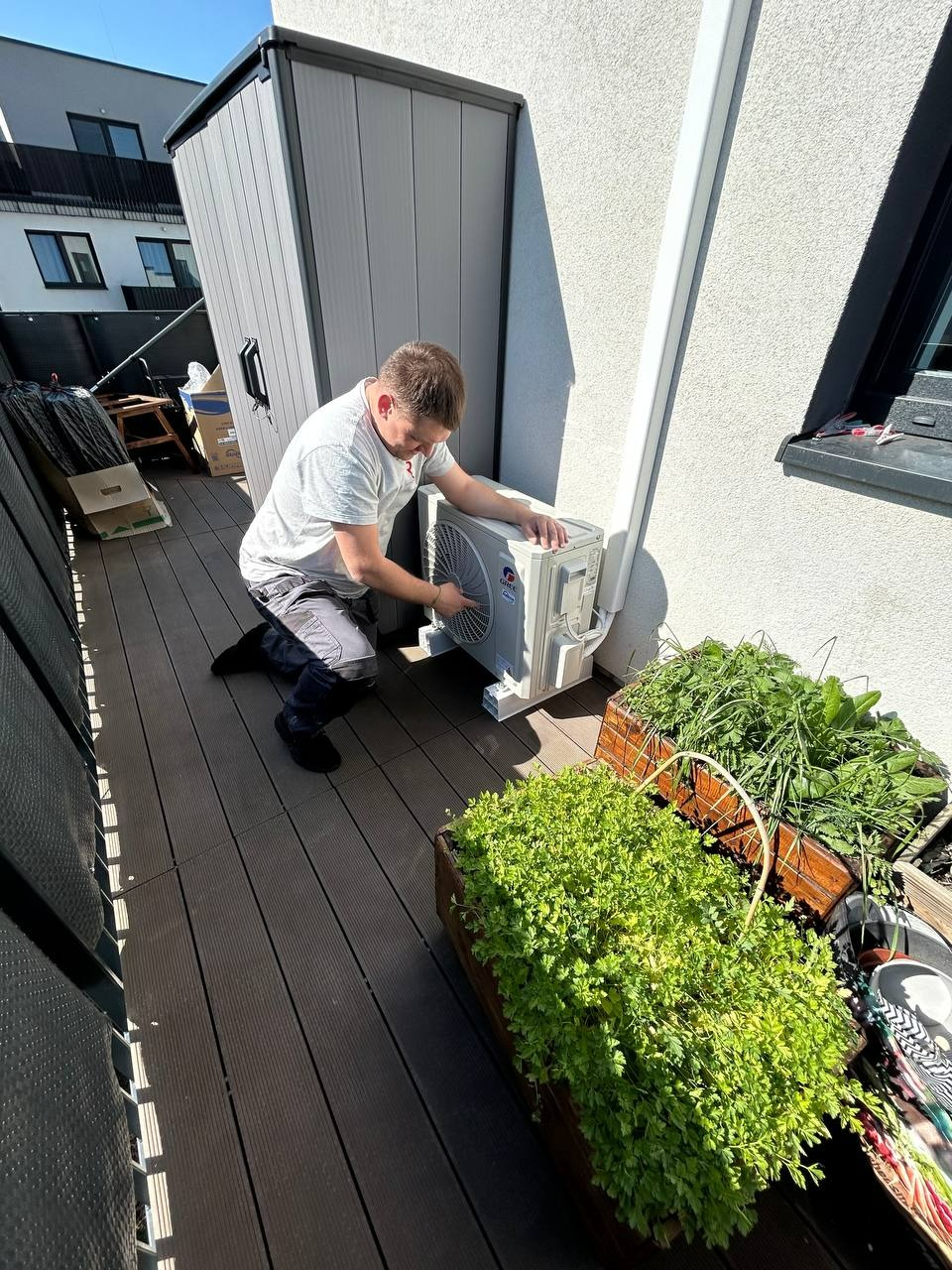 Instalacja klimatyzacji na balkonie we Wrocławiu. Mężczyzna w szarym stroju montuje biały klimatyzator. Obok skrzynki z zielenią i narzędzia.