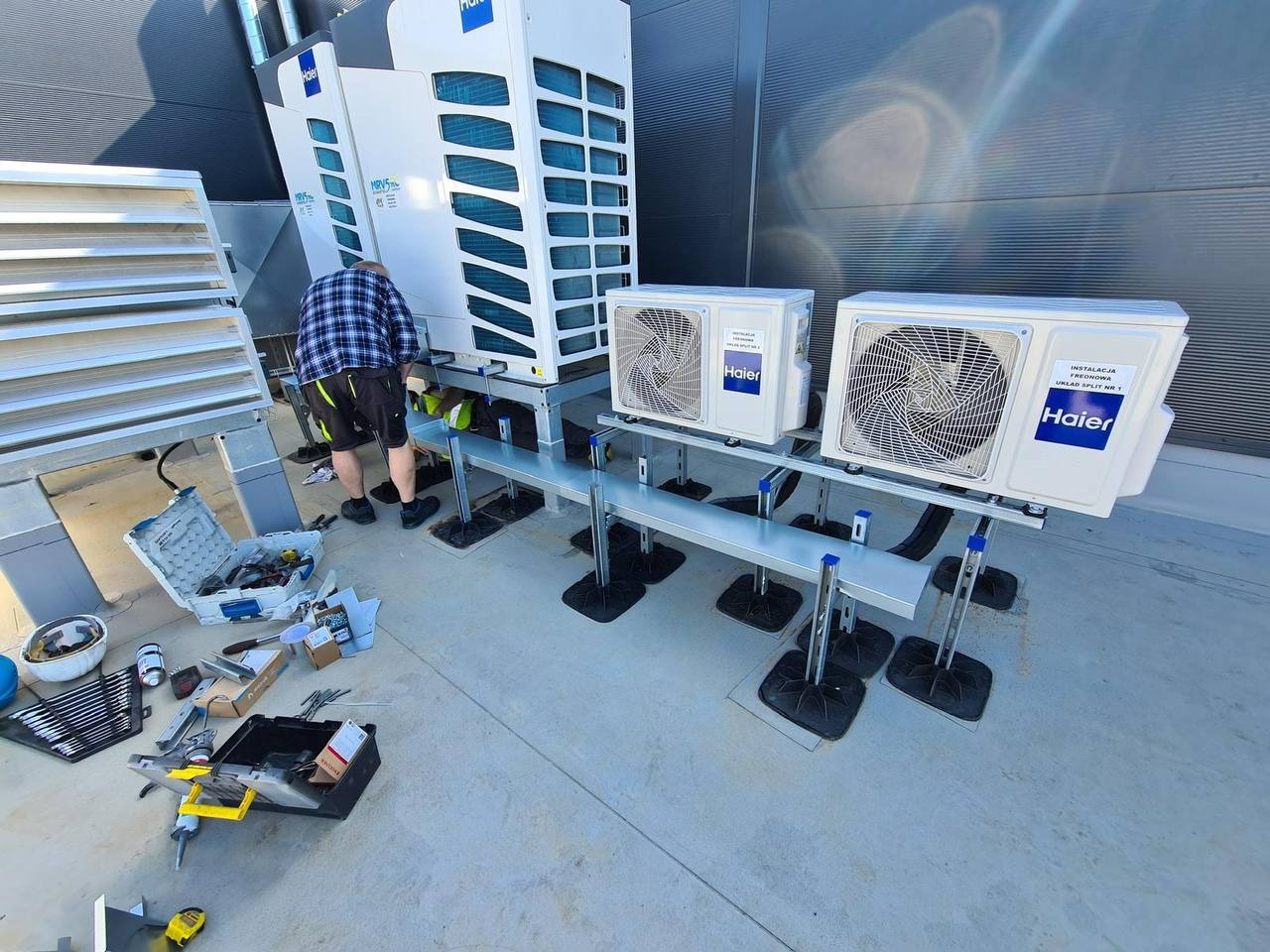 Montaż klimatyzacji Haier na dachu budynku. Widoczne jednostki zewnętrzne split, narzędzia pracy i instalator w trakcie podłączania. Szare tło dachu i elementy wentylacji.