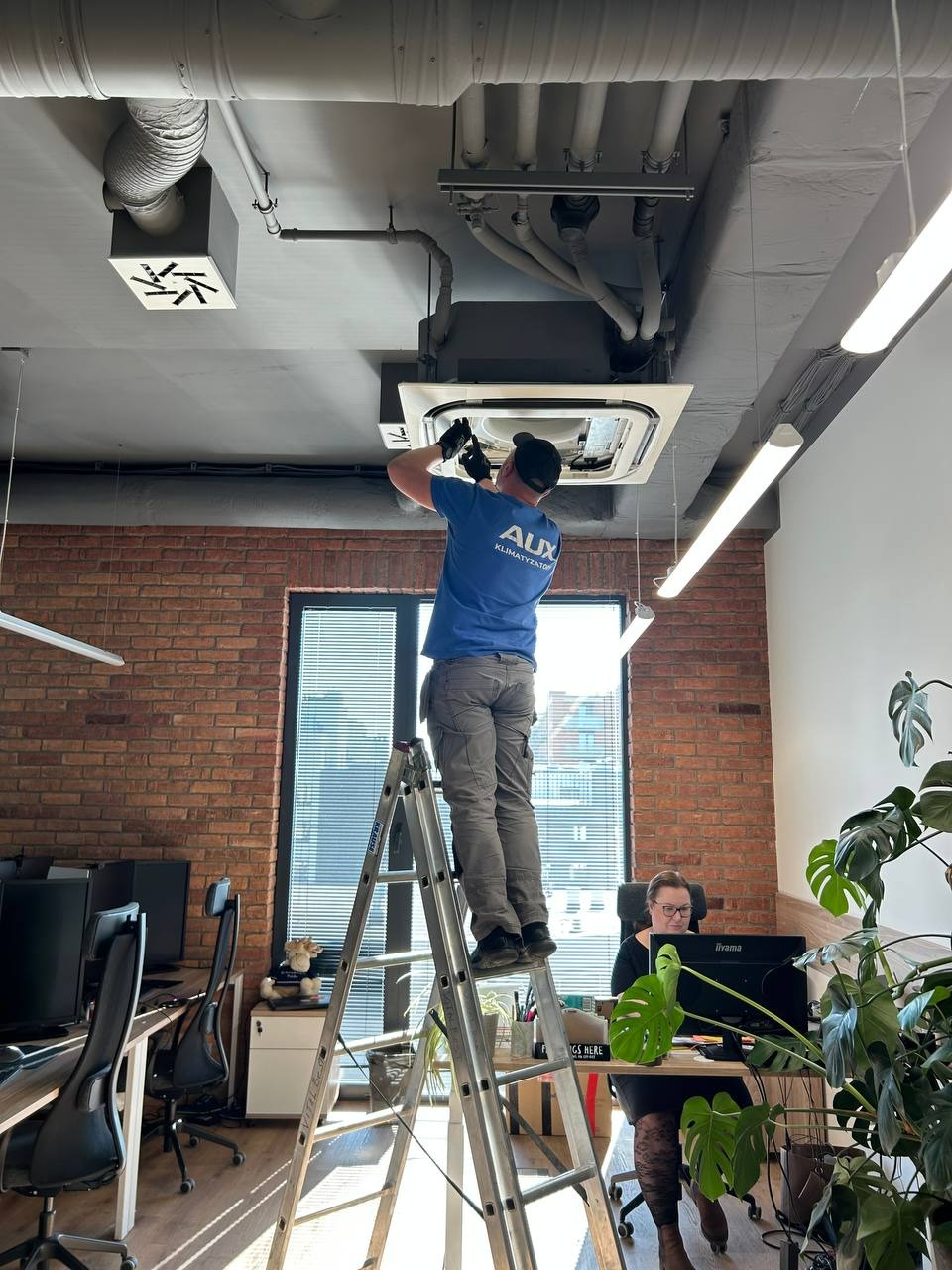 Mężczyzna na drabinie, w firmowej koszulce AUX, instaluje klimatyzację kasetonową w biurze z ceglaną ścianą. Widoczny pracownik biurowy i rośliny doniczkowe.